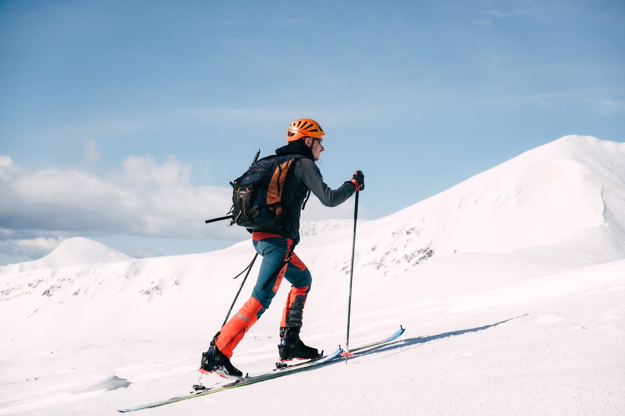 Skier climbing a snowy mountain in bright attire with poles, under a clear blue sky.